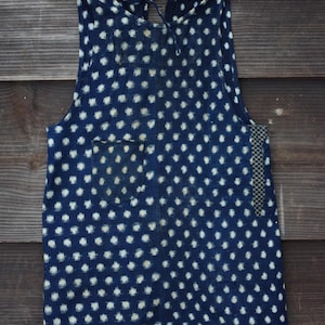 以下が含まれることがあります： インディゴブルーのノースリーブトップス。白い水玉模様が特徴です。小さなポケットと、側面にチェック柄のアクセントがあります。首元は紐で結ばれています。