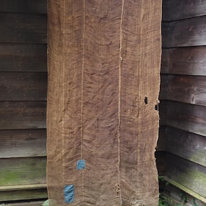 Op de afbeelding: Een groot, rechthoekig stuk bruine, grof geweven stof dat verticaal hangt. Het textiel heeft zichtbare verticale naden, een klein gat aan de rechterkant en twee kleine, rechthoekige blauwe lapjes die aan de linkeronderkant zijn vastgenaaid.