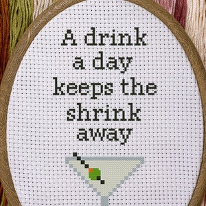 May include: A white embroidery hoop with a brown wooden frame. The embroidery is a cross-stitch design of a martini glass with an olive and the text "A drink a day keeps the shrink away".