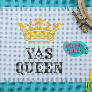 May include: A white cross-stitch pattern with a yellow crown and the text "YAS QUEEN". The pattern is on a white Aida cloth and is surrounded by colorful embroidery floss.
