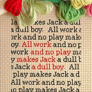 May include: A close-up of a cross-stitch sampler with the text "All work and no play makes Jack a dull boy." The sampler is made with colorful embroidery floss on a beige linen fabric.