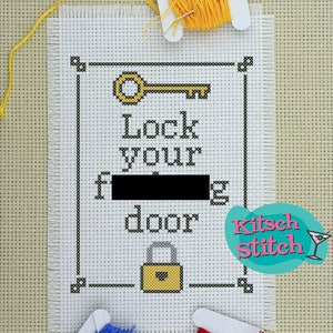 Peut inclure: Une broderie au point de croix avec le texte "Lock your f***ing door" et un motif de clé et de serrure. La pièce est encadrée de points noirs et comprend des bobines de fil jaune, bleu et rouge. Un logo "Kitsch Stitch" est dans le coin.