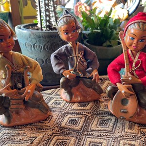 May include: Three vintage ceramic figurines of musicians. Each figure is seated and playing a different instrument. The figures wear colorful clothing and head coverings. The figurines are on a patterned surface, with plants in the background.