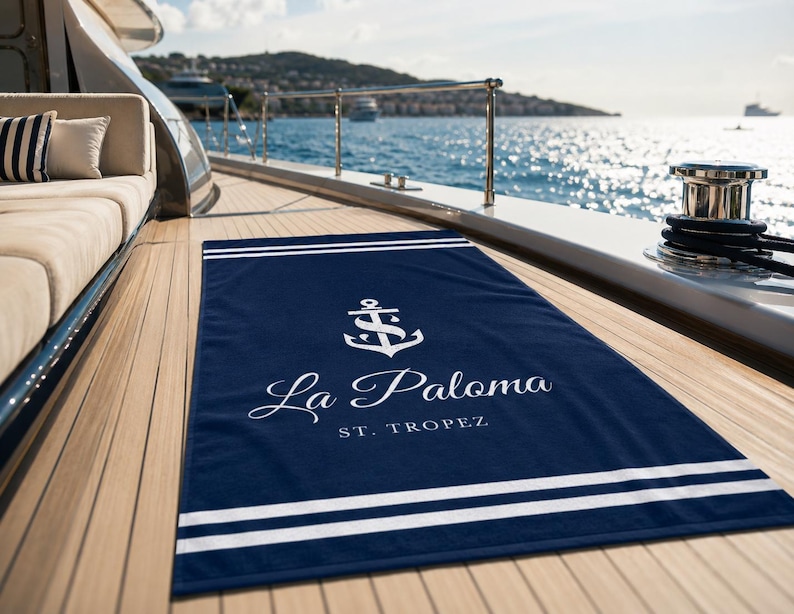 Könnte beinhalten: Marineblaues Strandtuch mit weißen Streifen und einem Ankerlogo. Der Text "La Paloma St. Tropez" ist in Weiß gehalten. Das Handtuch liegt auf einem Holzdeck mit Blick auf das Meer.