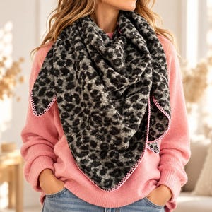 Könnte beinhalten: Ein dreieckiges Tuch mit schwarz-grauem Leopardenmuster und einem leuchtend rosa genähten Rand, getragen über einem einfarbigen rosa Langarmpullover und einer blauen Jeans. Das Tuch hat eine strukturierte Optik und einen markanten Saum.