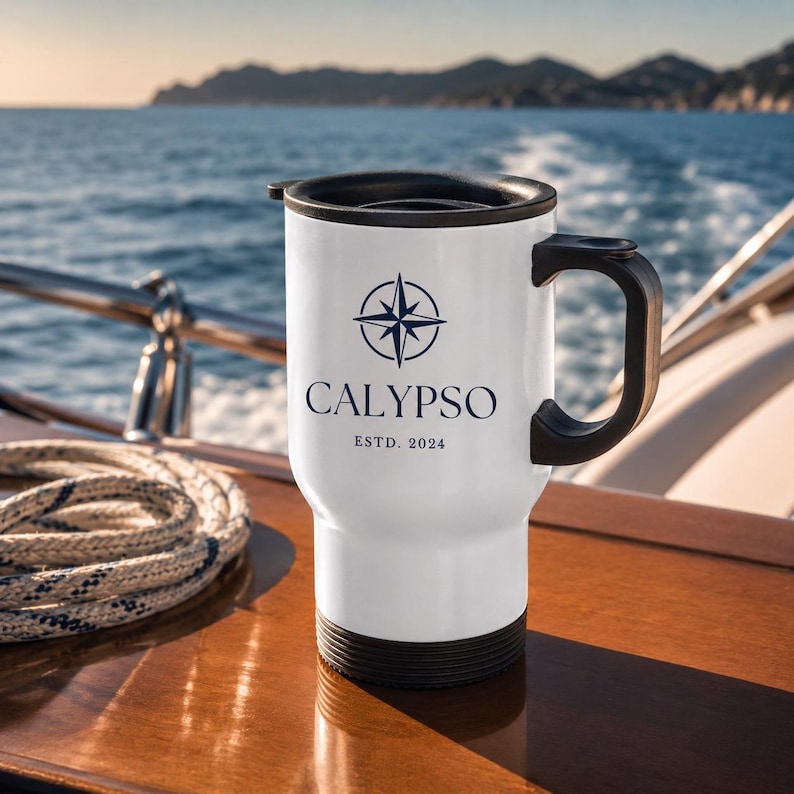 Könnte beinhalten: Weißer Reisebecher mit schwarzem Griff und Deckel. Der Becher trägt das Wort "CALYPSO" in Marineblau, zusammen mit einer Kompassrosen-Grafik und dem Text "ESTD. 2024". Der Becher steht auf einer Holzoberfläche mit einem Seil und dem Ozean im Hintergrund.