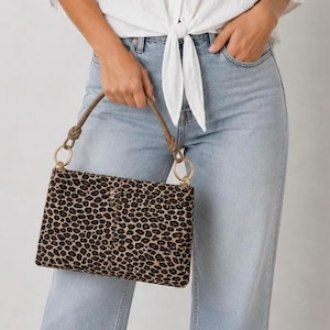 Könnte beinhalten: Eine rechteckige Handtasche mit Leopardenmuster in Braun- und Beigetönen. Die Tasche hat einen braunen Griff mit goldfarbenen Ringen und einem geknoteten Detail. Die Tasche wird von einer Person gehalten, die eine hellblaue Jeans und ein weißes Hemd trägt.