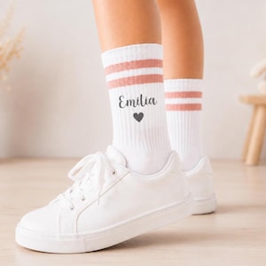Könnte beinhalten: Weiße Crew-Socken mit zwei horizontalen altrosa Streifen am oberen Rand. Der Schriftzug "Emilia" ist in einer dunkelgrauen Schreibschrift über einem kleinen dunkelgrauen Herzsymbol aufgedruckt. Die Socken werden mit weißen Sneakern getragen.