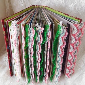Op de afbeelding: Een handgemaakt dagboek met een rode, groene en witte stoffen omslag. De pagina's zijn versierd met kant in verschillende kleuren, waaronder wit, roze en groen.