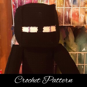 May include: Crochet pattern for a black Minecraft character with white and purple eyes. The character is standing in front of a white wire shelf with colourful items on it.