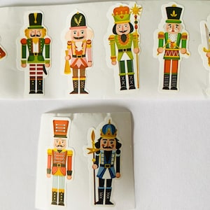 May include: A collection of eight die-cut stickers featuring nutcracker figures in various uniforms. The characters wear colourful hats, jackets, and boots, with some holding instruments, staffs, or swords. The stickers are arranged on two white sheets.