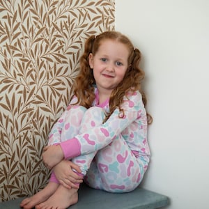May include: A young person with red curly hair in pigtails wearing patterned pajamas. The pajamas have a pink, blue, and white design. The person is sitting on a blue bench in front of a wall with a brown and white leaf pattern.