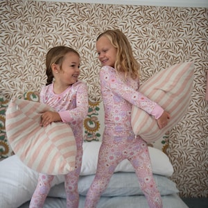 May include: Two young people in matching pink floral pajamas stand on a bed, each holding a striped pillow. The pajamas have a floral pattern, and the pillows have pink and white stripes. The background features a patterned wallpaper.
