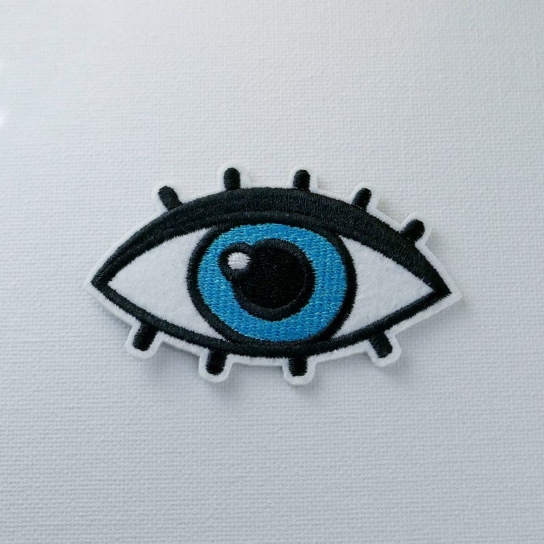 May include: A blue eye with black eyelashes and a white pupil, embroidered on a white fabric.