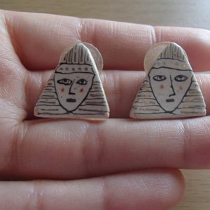 May include: A pair of handmade ceramic stud earrings. Each earring is a white triangle with a stylized face design. The face has black outlines, red cheeks, and gold accents. The earrings are small, likely less than 1 inch in size.