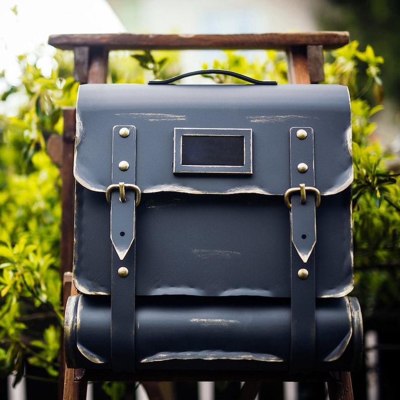 May include: A dark blue metal mailbox with a distressed finish and gold accents. The mailbox has a rectangular opening with a black metal flap and a handle on the top.