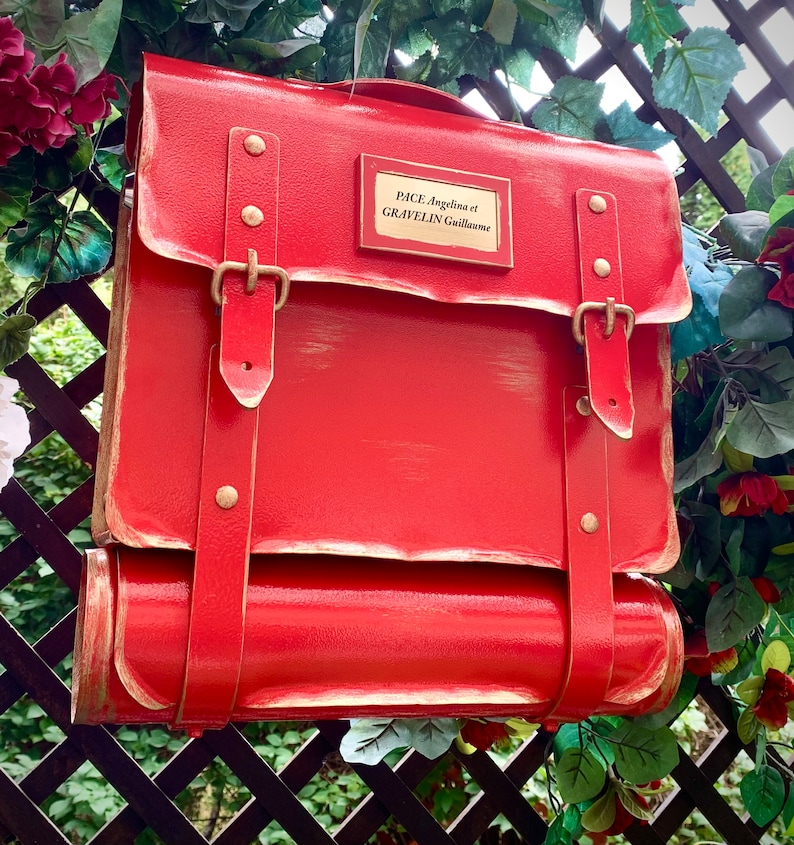 May include: A red metal mailbox with a brass nameplate that reads "PACE Angelina et GRAVELIN Guillaume". The mailbox has a rolled top and two straps with brass buckles.