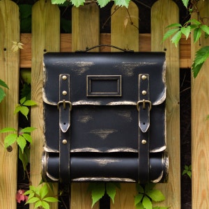 May include: Black metal mailbox with gold accents and a distressed finish. The mailbox has two straps and a roll-top design.