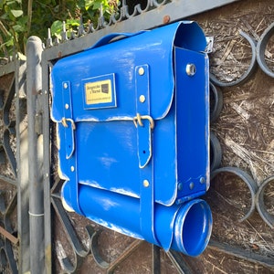 May include: A blue mailbox shaped like a vintage briefcase with a gold nameplate that reads "Skrzynka i Normal" and a website address. The mailbox has a flap that opens for mail delivery.