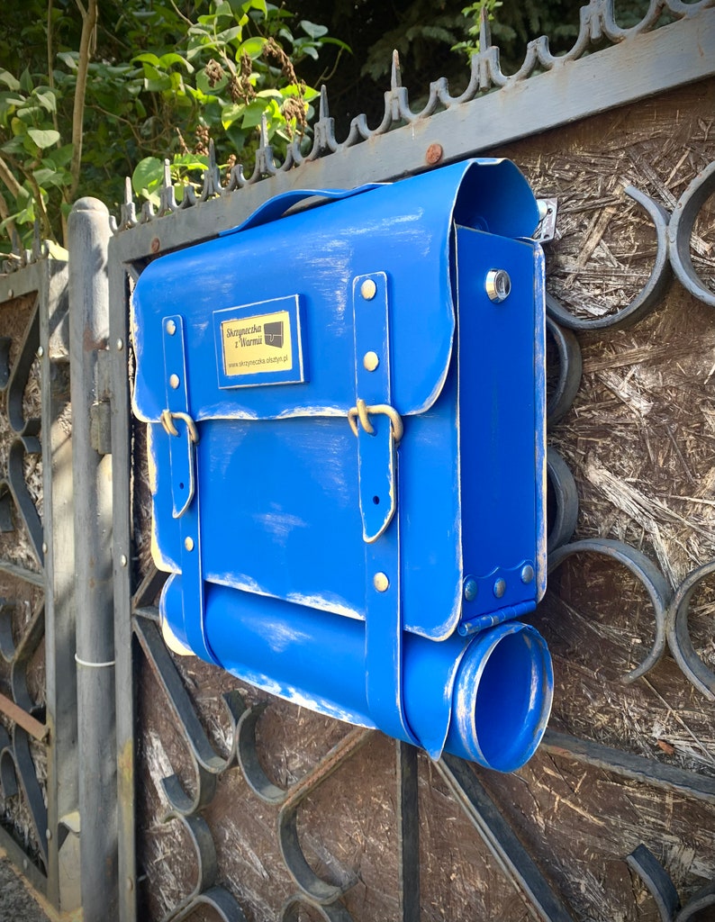 May include: A blue mailbox shaped like a vintage briefcase with a gold nameplate that reads "Skrzynka i Normal" and a website address. The mailbox has a flap that opens for mail delivery.