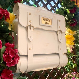 May include: White painted mailbox with a gold plaque that reads "Familie Kaun". The mailbox has two straps and a rolled top.