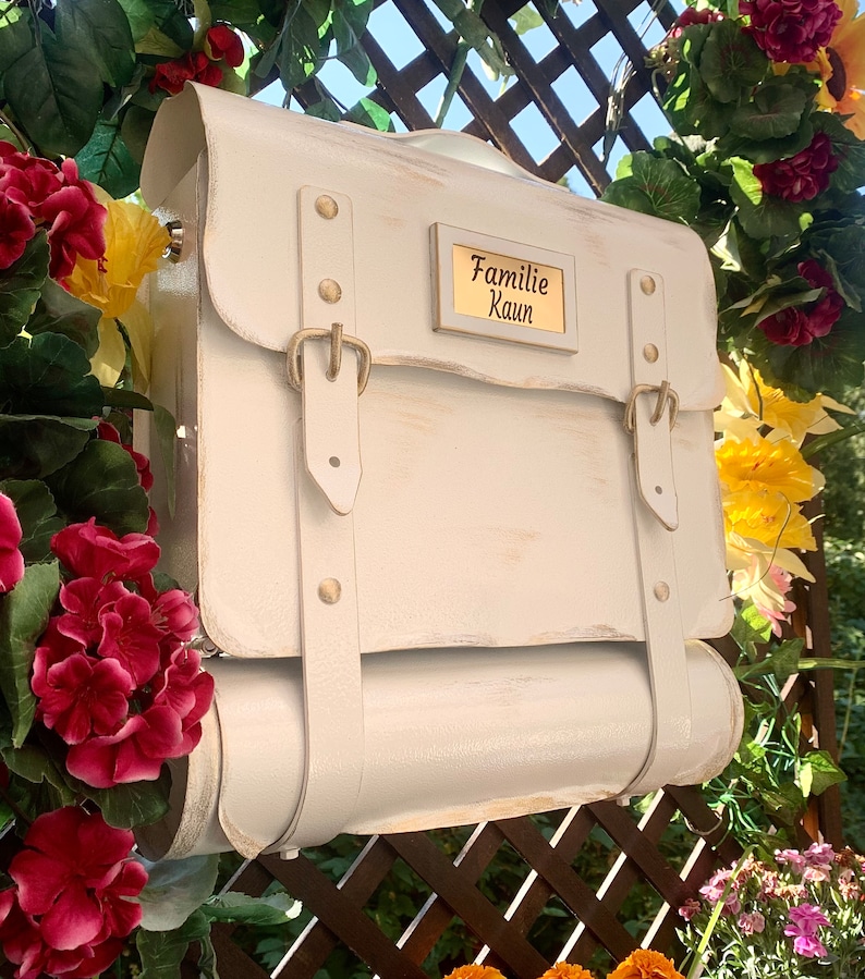 May include: White painted mailbox with a gold plaque that reads "Familie Kaun". The mailbox has two straps and a rolled top.