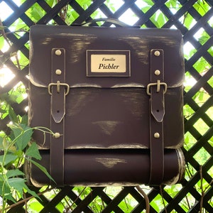 May include: A brown metal mailbox with a distressed finish and gold accents. It has two leather straps with buckles and a brass nameplate that reads "Familie Pichler".