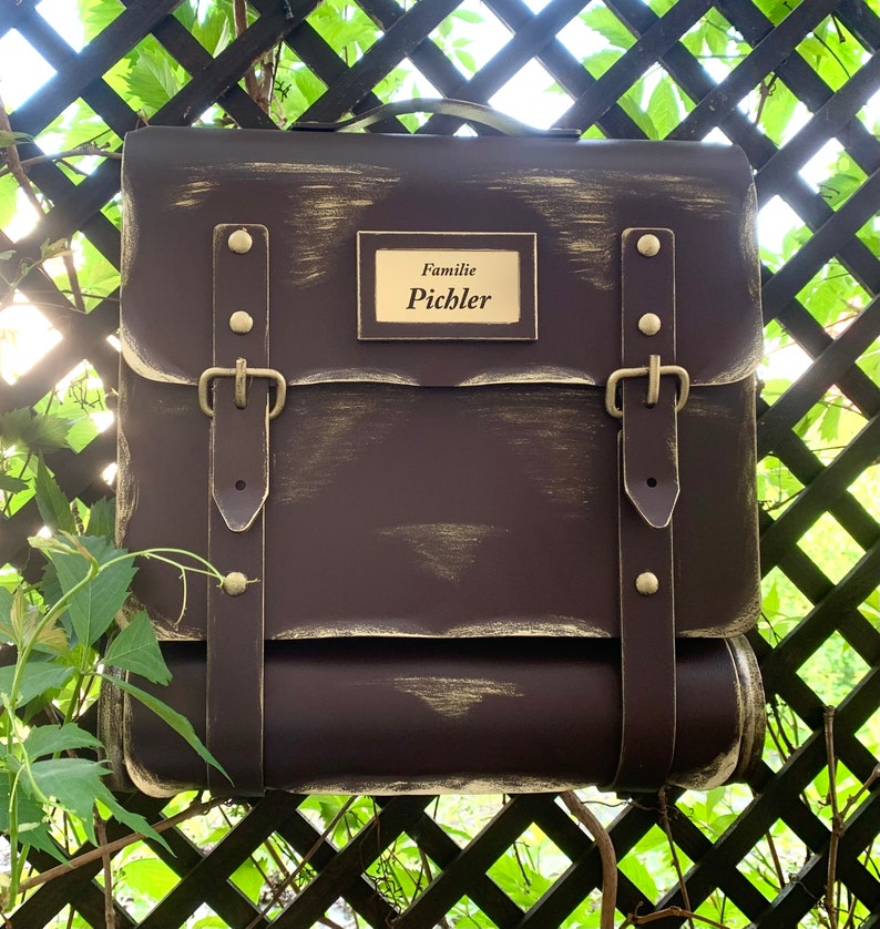 May include: A brown metal mailbox with a distressed finish and gold accents. It has two leather straps with buckles and a brass nameplate that reads "Familie Pichler".
