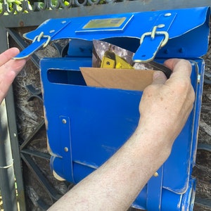 May include: A blue metal mailbox with a flap open, revealing a brown envelope and a postcard. The mailbox has a vintage design with leather straps and buckles.