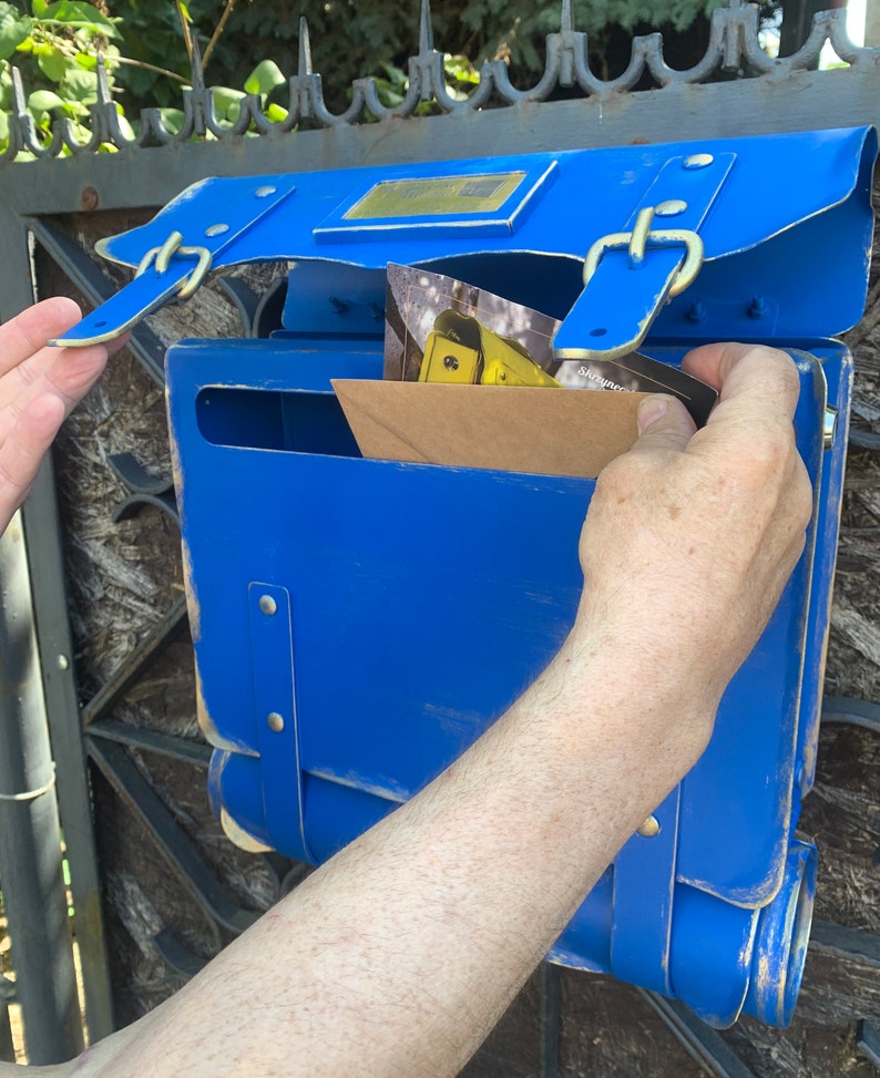 May include: A blue metal mailbox with a flap open, revealing a brown envelope and a postcard. The mailbox has a vintage design with leather straps and buckles.