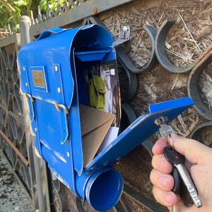 May include: A blue metal mailbox shaped like a backpack with a keyhole and a key in the lock. The mailbox has a flap that is open and a tube for mail.