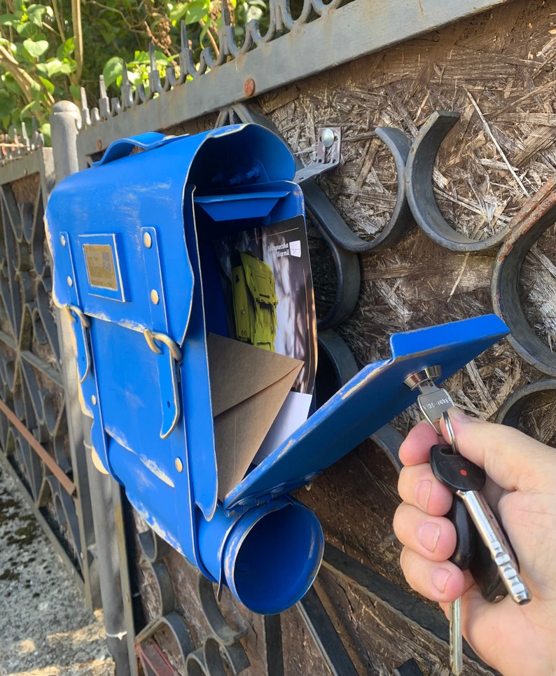 May include: A blue metal mailbox shaped like a backpack with a keyhole and a key in the lock. The mailbox has a flap that is open and a tube for mail.