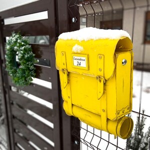 May include: A yellow mailbox shaped like a vintage school bag with a metal flap and a snow-covered top. The mailbox has a label that reads "Lawendowa 24".