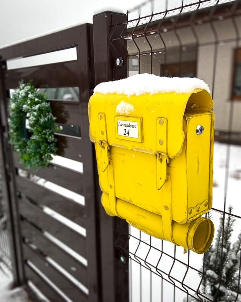 May include: A yellow mailbox shaped like a vintage school bag with a metal flap and a snow-covered top. The mailbox has a label that reads "Lawendowa 24".