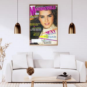 Puede incluir: Una portada de revista vintage enmarcada con el título "Mademoiselle" y una foto de una mujer. La revista trata sobre moda y belleza. El marco está encima de un sofá blanco en un salón moderno.