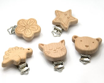May include: Five wooden baby pacifier clips in various shapes: a flower, a star, a dinosaur, a cat, and a bear. Each clip has a silver metal clasp for attaching to clothing. The wooden shapes are light brown with engraved details.