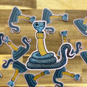 Peut inclure: Un autocollant découpé représentant un serpent bleu enroulé à la base, avec une manche de pull en tricot jaune recouvrant son cou et sa tête. Le serpent présente un motif d'écailles détaillé et une tête bleu clair. Le motif est répété.