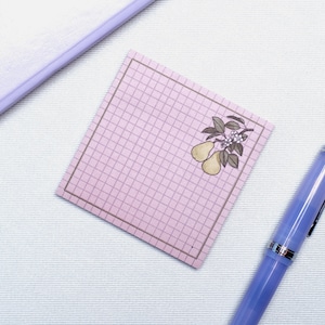 Peut inclure: Un bloc-notes carré rose avec un motif quadrillé et une bordure. Le coin supérieur droit présente une illustration botanique de poires et de feuilles. Un stylo et un carnet lilas sont également dans l'image.