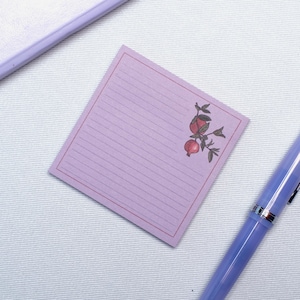 May include: A lilac notepad with lined pages and a decorative border. A botanical illustration of pomegranates and leaves is in the upper right corner. A matching lilac pen is in the foreground, with a glimpse of a lilac notebook in the background.