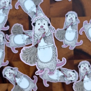 May include: A die-cut sticker featuring a detailed illustration of an opossum dressed as a maid. The opossum has white and grey fur, pink ears, and a pink nose. It wears a white ruffled apron and cap while holding a brown straw broom.