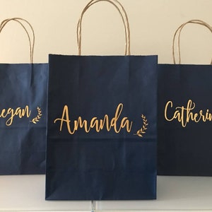 May include: Three navy blue gift bags with brown paper handles. Each bag has a name in gold script: "Megan", "Amanda", and "Catherine". A gold leaf design is next to each name.