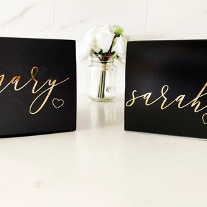 May include: Two black gift boxes with gold lettering. The box on the left says "mary" with a small gold heart. The box on the right says "sarah" with a small gold heart.