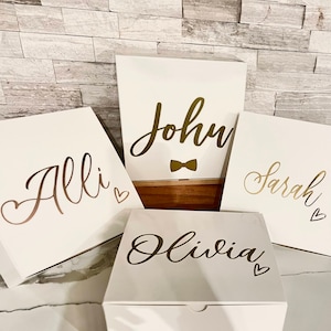 May include: Four white gift boxes with gold lettering. The boxes are labeled "Allie", "John", "Olivia", and "Sarah". The box labeled "John" also has a gold bow tie.