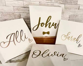 May include: Four white gift boxes with gold lettering. The boxes are labeled "Allie", "John", "Olivia", and "Sarah". The box labeled "John" also has a gold bow tie.