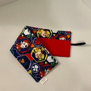 May include: A blue and red fabric pencil roll with a Paw Patrol theme. The roll has multiple pockets for storing pencils and other art supplies. The roll is closed with a black strap.