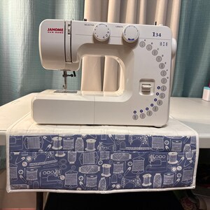 May include: A white Janome New Home 134 sewing machine with two circular control dials and a vertical stitch selection panel. The machine sits on a quilted mat featuring a blue and white pattern of sewing supplies like spools and needles.