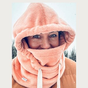 May include: A close-up of a person wearing a peach-colored fleece hooded neck warmer with a white drawstring. The neck warmer is pulled up over the person's face, covering their nose and mouth. The person's eyes are visible and they are looking directly at the camera. The background is blurry and appears to be a snowy landscape.