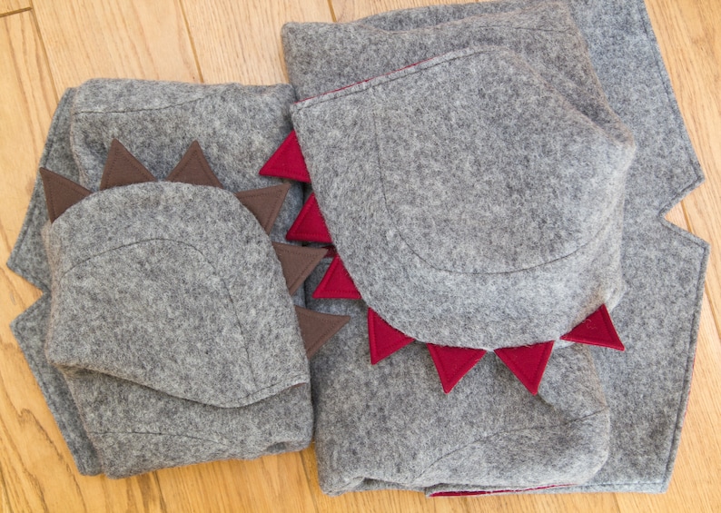 May include: Two gray felt dinosaur costumes with red and brown felt accents. The costumes have a hood with a pointed shape and a tail.