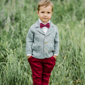 Puede incluir: Un ni&ntilde;o peque&ntilde;o con un blazer de lino gris, una pajarita roja, pantalones rojos y zapatos negros. Est&aacute; de pie sobre una superficie de madera con hierba verde en el fondo.