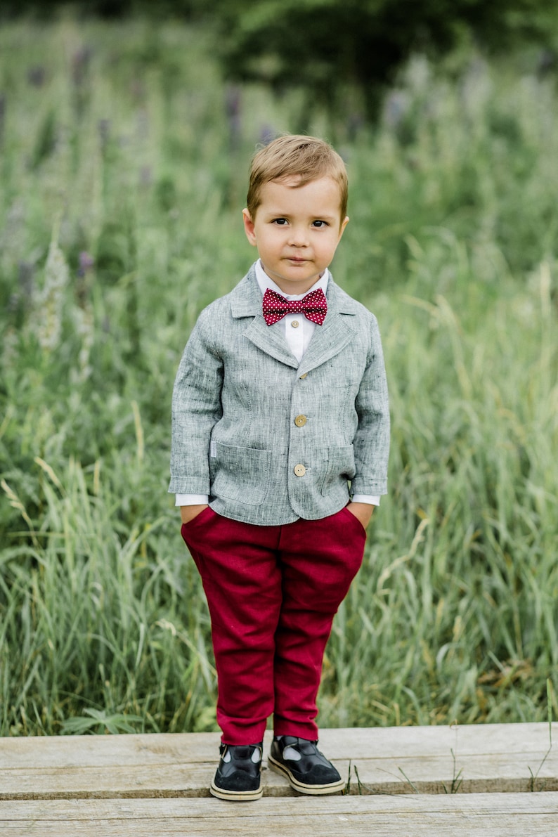 Puede incluir: Un ni&ntilde;o peque&ntilde;o con un blazer de lino gris, una pajarita roja, pantalones rojos y zapatos negros. Est&aacute; de pie sobre una superficie de madera con hierba verde en el fondo.